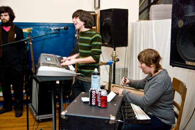 Drei Männer, einer steht links, ein anderer spielt Piano und singt, und der dritte singt, während er Getränkedosen auf einem Tisch hält, mit einem Lautsprecher und einem Vorhang im Hintergrund.
