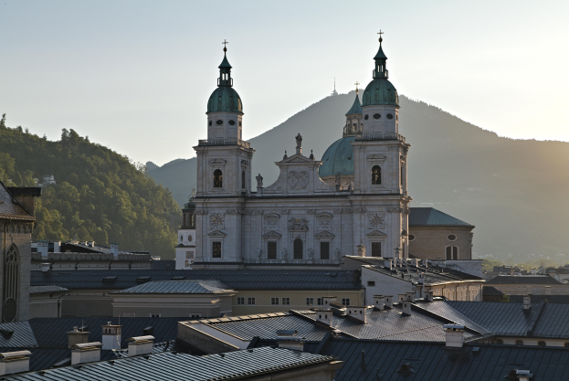 Eine Panoramasicht auf Salzburg, Österreich von einem Hügel aus, mit Gebäuden, Bäumen, einer prominenten Kathedrale, einem blauen und weißen Himmel und fernen Bergen.