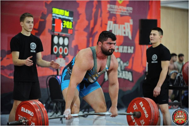 Ein Mann hebt eine Hantel bei einem Gewichtheben-Wettbewerb, umgeben von Zuschauern, mit Wettbewerbsequipment und Signage, die die 2018 World Weightlifting Championships anzeigen.