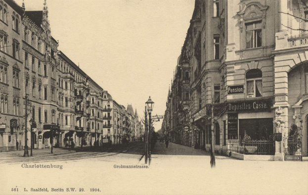 Schwarzes und weißes Foto einer Straßenansicht in Charlottenburg, Deutschland, mit Gebäuden, Straßenlaternen, Schildern, Motorfahrzeugen, Fußgängern, Gittern und einem klaren Himmel.