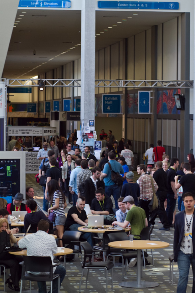 Ein belebter Flughafen-Foyer mit wartenden Menschen, einige stehen und andere arbeiten, Richtungsschilder an der rechten Wand und oben, und Lampen, die von der Decke hängen.