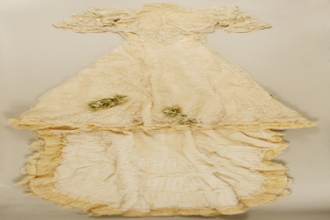 Weißes Brautkleid mit Rüschen und floraler Stickerei, auf einem weißen Hintergrund präsentiert, mit einer taillierten Taille, Herzausschnitt, Spitzenverzierung und einem langen, üppigen Rock.