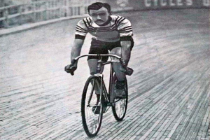 Ein Schwarz-Weiß-Foto eines Mannes, der auf einer Rennbahn Fahrrad fährt, mit einem Zaun und einer Tafel im Hintergrund, der angeblich Sir Ernest Hemingway ist.