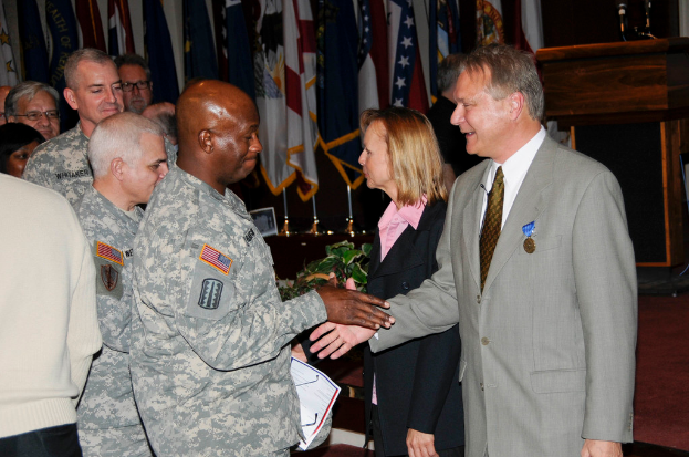 Eine Gruppe von Menschen mit zwei Personen, die Hände schütteln, Flaggen im Hintergrund, ein Podium mit Mikrofon und eine Topfpflanze in der Nähe.