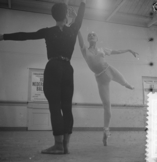 Ein Schwarz-Weiß-Foto von einem Mann und einer Frau, die in einem Studio tanzen, wobei die Frau in der Luft ist und Plakate an der Wand hinter ihnen hängen, die von Lichtern an der Oberseite des Bildes beleuchtet werden.