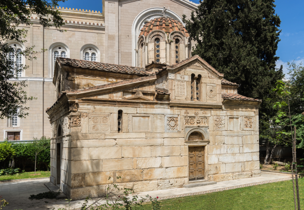 Die Kirche von St. Nikolaos in Thessaloniki, Griechenland, ein Gebäude mit Fenstern und einer Tür, umgeben von Pflanzen, Gras, Bäumen und unter einem bewölktem Himmel.