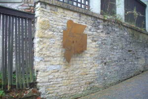 Steinmauer mit Metalltor auf der linken Seite, eine Gedenktafel für die Eröffnung des alten Rathauses und ein Gebäude mit Fenstern und Pflanzen im Hintergrund.