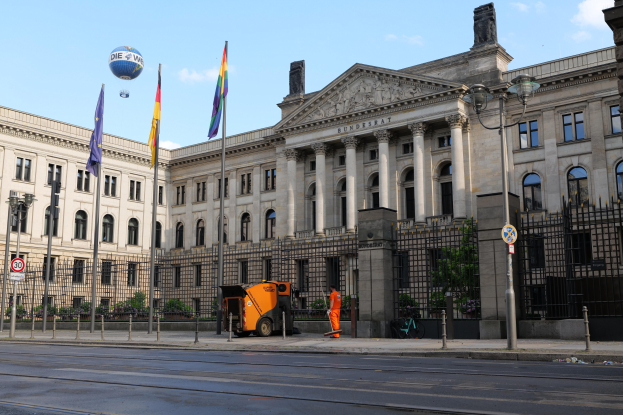 Großes modernes Gebäude mit zahlreichen Fenstern, identifiziert als der Bundestag in Berlin, Deutschland, umgeben von Straßeninfrastruktur, Fahrzeugen, Passanten und Bäumen unter einem bewölktem Himmel.
