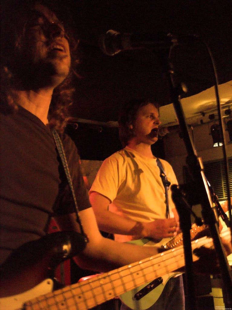 Eine Person in einem weißen Hemd singt in ein Mikrofon und spielt eine grüne Gitarre, während sie neben einer anderen Person in einem schwarzen Hemd und Brille steht, die ebenfalls singt und eine schwarze Gitarre spielt, mit Scheinwerfern und anderen Instrumenten im Hintergrund.