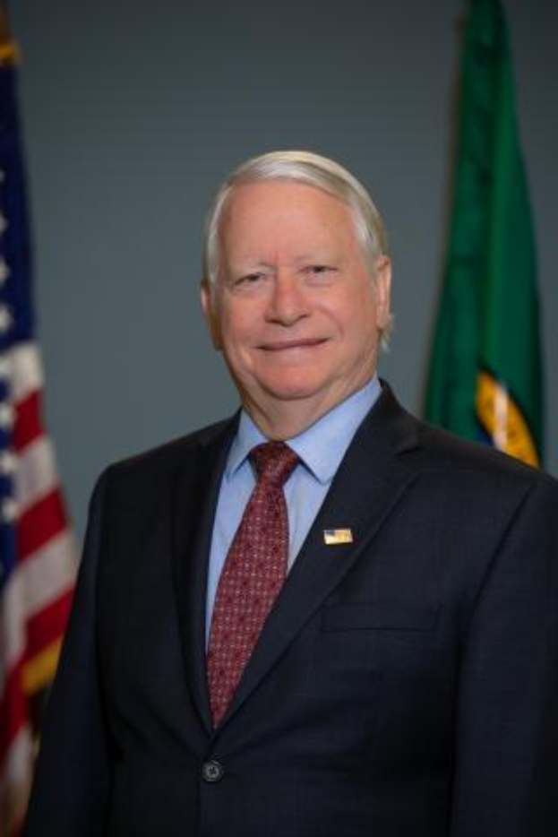 Ein Mann im Anzug und Krawatte steht lächelnd vor einer amerikanischen Flagge, mit einer Wand im Hintergrund.