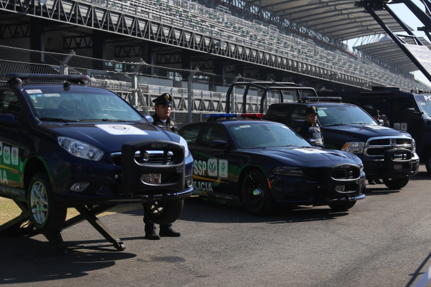 Polizeiautos vor einer Rennstrecke mit uniformierten Personen auf der Straße, im Hintergrund ein Stadion und eine Absperrung.