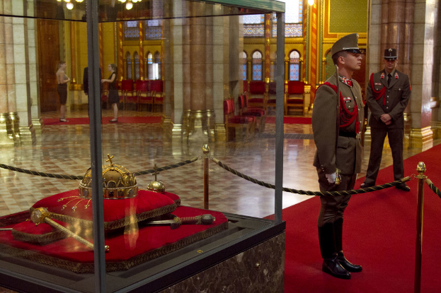 Fünf Personen stehen auf Teppichen mit Stangen, Seilen und Gegenständen in einer durchsichtigen Glasbox links, eine Säule, Licht, Stühle und eine Wand dahinter und Kronleuchter über drei der Personen.