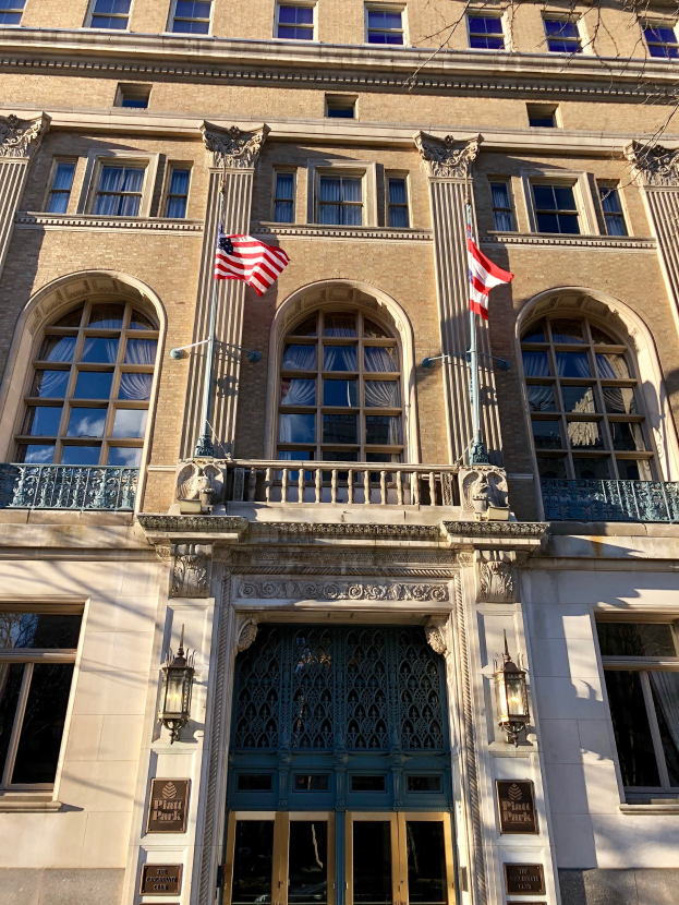 Außenansicht des Plaza Hotels in New York City, mit Glasfenstern, -türen, -säulen, -geländern, -leuchten, -schildern und -flaggen vor einem Himmelhintergrund.