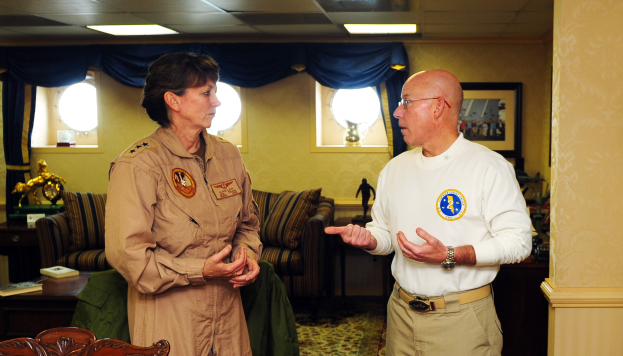 Ein Mann und eine Frau unterhalten sich in einem Wohnzimmer mit Möbeln, Bücherregalen und einem Foto auf dem Wand, mit Fenstern und Vorhängen im Hintergrund und einer Decke mit Lampen.