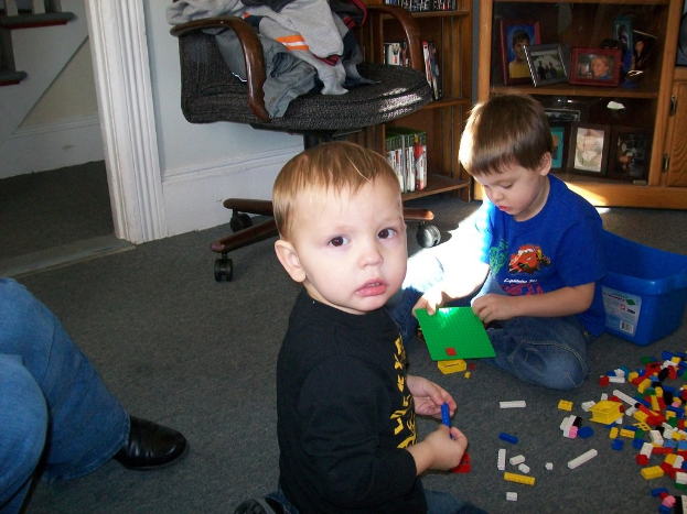 Ein kleiner Junge steht in der Mitte des Bildes, ein anderer Junge liegt rechts auf dem Boden, umgeben von Spielzeugblöcken, mit Rissen an der Wand, einem Stuhl mit Kleidung in der Mitte, einer Treppe oben rechts und einem Mann links.