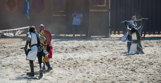 Eine Person steht in der Mitte und h├Ąlt eine Kamera, um Bilder von einem Kampf zwischen zwei Gruppen auf beiden Seiten zu machen, mit einer Wand, einem Tor und einer Flagge im Hintergrund und Sand unten.