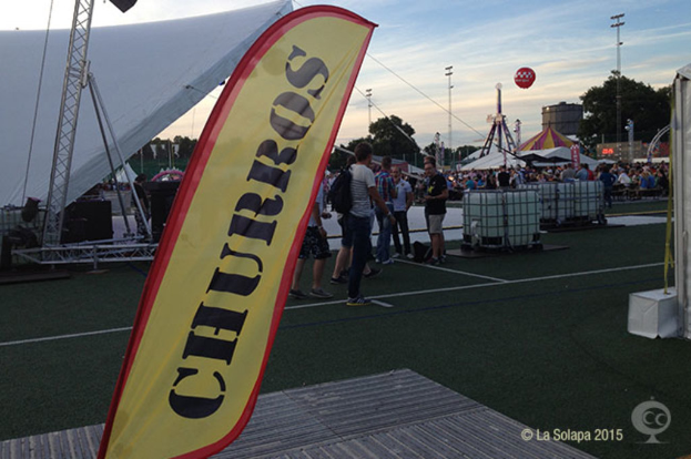 Eine Karnevalsszene mit Zelten, einem zentralen Mast, Menschen, Bäumen, Strommasten, Straßenbahnen auf der Straße und einem Eisenständer auf der linken Seite unter einem Himmel.