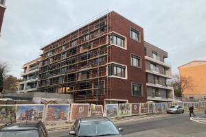 Großes Gebäude mit Gerüst an der Seite, umgeben von Bäumen unter einem klaren blauen Himmel, mit Fahrzeugen auf der Straße und einer Person auf dem Bürgersteig, was auf laufende Bauarbeiten hinweist.