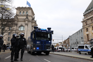 Eine Gruppe von Polizisten in Helmen und bewaffnet mit Gewehren steht vor einem Gebäude mit Fenstern, Säulen, Bögen und einer Flagge oben drauf, mit Fahrzeugen auf der Straße, Bäumen, Laternenmasten und einem bewölkten Himmel im Hintergrund.