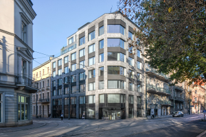 Großes Bürogebäude mit zahlreichen Fenstern, Straßeninfrastruktur einschließlich Masten und Kabeln, Fahrzeuge auf der Straße, Fußgänger auf dem Gehweg, Bäume und bewölkter Himmel, das als neue Firmenzentrale dient.