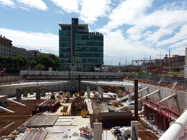 Eine Baustelle mit einem großen Gebäude, das wahrscheinlich der Europäische Union Hauptsitz ist, umgeben von Bäumen, Pfählen und Lichtern unter einem bewölkten Himmel.