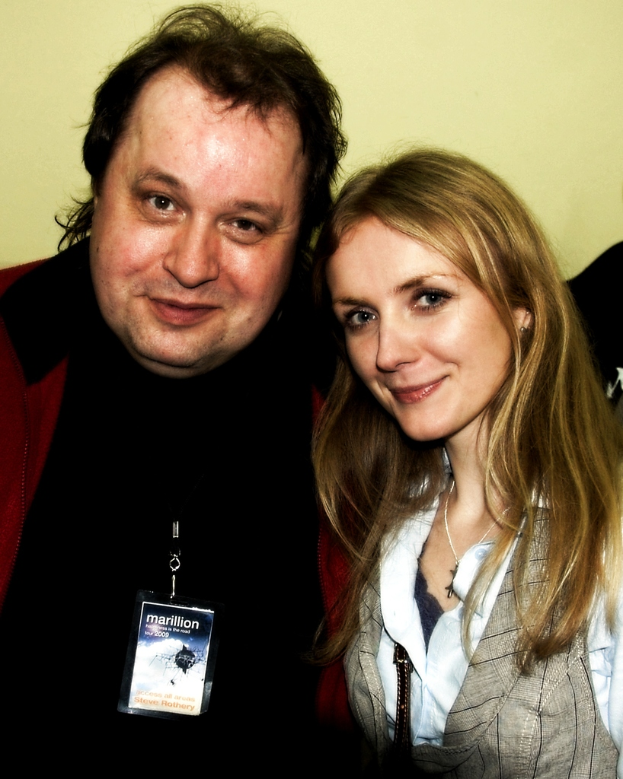 Eine Frau sitzt auf einem Stuhl und trägt eine schwarze Jacke und ein gelbes Kleid, an ihrer Kleidung ist ein Abzeichen befestigt, vor einer Wand.