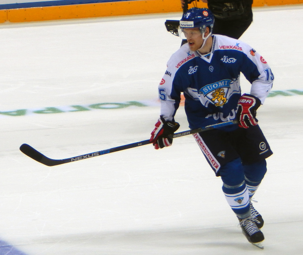 Ein Eishockeyspieler in Aktion auf dem Eis, der einen Helm, Handschuhe und Schlittschuhschuhe trägt und einen Eishockeyschläger hält, während eine andere Person eine Kamera und eine Wand mit Text im Hintergrund hält.