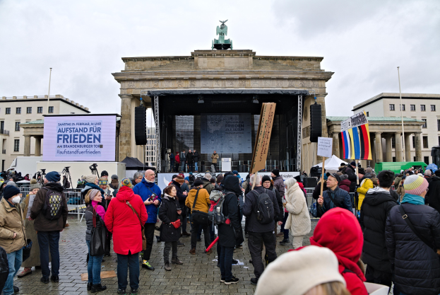 Menschenmenge versammelt sich vor einem Gebäude mit einer Bühne, auf der Redner und ein Bildschirm zu sehen sind, umgeben von Fahnen und Transparenten, in Berlin während einer Demonstration.
