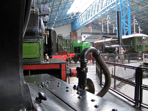 Mehrere Lokomotiven in einem Lagerhaus mit Eisenstangen oben, einem Zaun auf der rechten Seite, einem Zug auf dem Bahnsteig neben dem Zaun und zwei Personen in der Mitte neben dem Zaun.