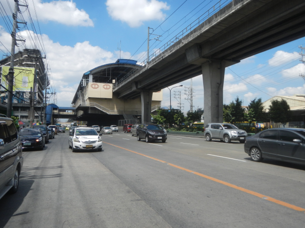 Vielbefahrene Straße mit fahrenden Autos, eine Brücke im Hintergrund, Strommasten mit Drähten, Bäume, Gebäude und ein bewölkter Himmel.