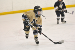 Zwei junge Jungs in Sportkleidung, Helmen, Handschuhen und Schlittschuhschuhen spielen Eishockey auf einem Eisplatz, jeder mit einem Hockeystock in der Hand, mit einer Wand im Hintergrund.