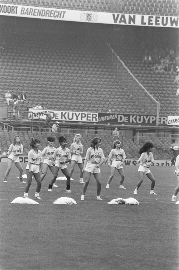 Eine Gruppe von Frauen auf einem Fußballfeld, umgeben von einer Menge im Publikum und Bannern mit Text, auf einem Schwarz-Weiß-Bild.