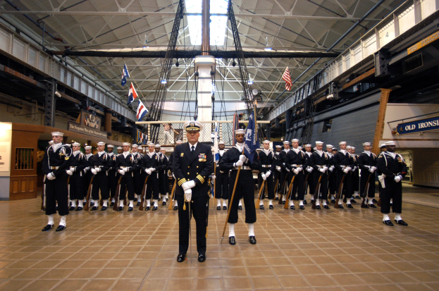 Gruppe uniformierter Männer in einem großen Raum mit Gewehren, die Kappen tragen und neben Fahnenmasten stehen, mit Texttafeln an den Wänden und Deckenleuchten.
