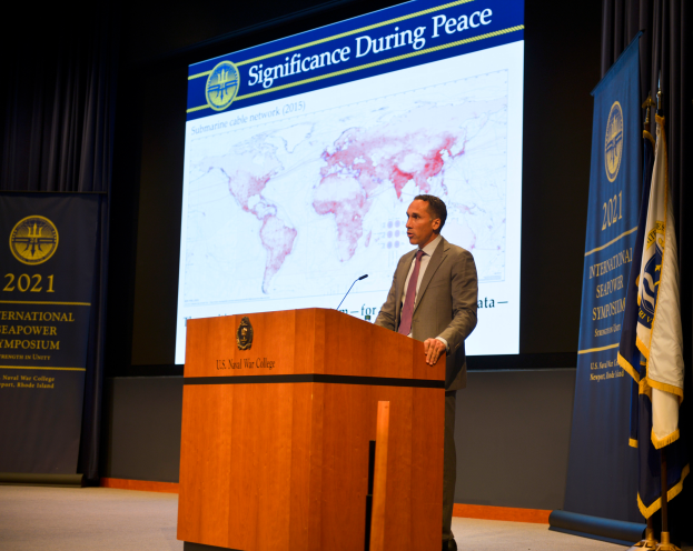 Ein Mann in Sakko, Krawatte und Hemd steht an einem Rednerpult mit einem Mikrofon, flankiert von zwei Fahnen und einem Banner mit Text und einer Karte im Hintergrund.