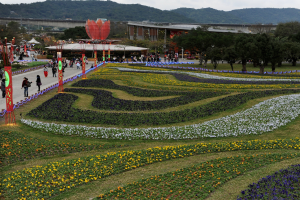 Großer Blumen Garten in einem Park mit Menschen, die herumlaufen, umgeben von grünem Gras, Bäumen, Gebäuden, einer Brücke, Bergen und einem klaren blauen Himmel, mit Pfählen und Lichtern auf der linken Seite.