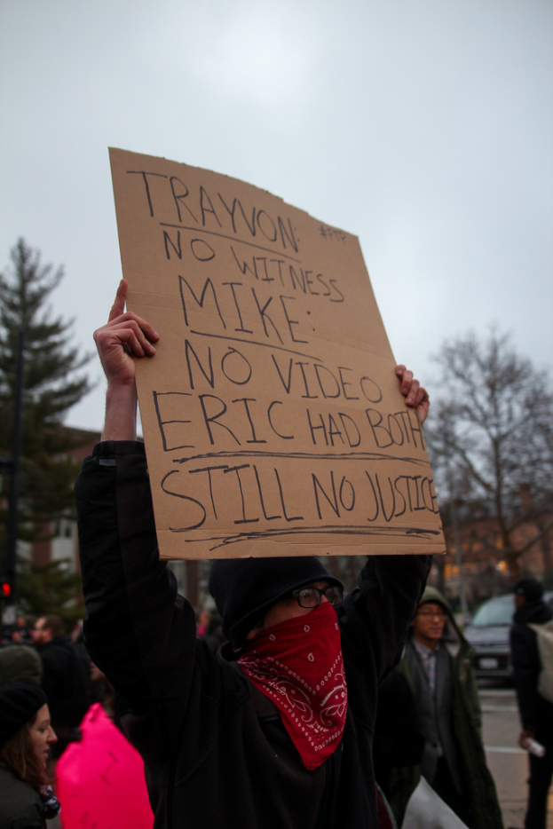 Eine Gruppe von Menschen, die auf der Straße protestieren, ein Mann hält ein Schild mit Text, mit Bäumen, einem Auto und dem Himmel im Hintergrund.