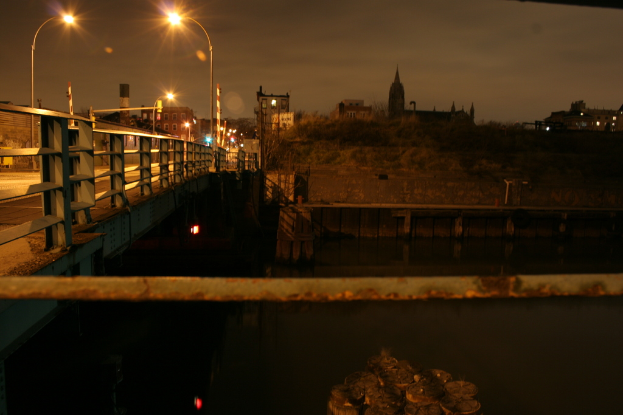 Eine Brücke überspannt einen Fluss mit zahlreichen Straßenlaternen darauf und im Hintergrund sind Gebäude zu sehen.