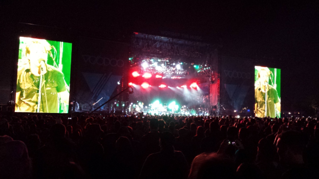 Konzertszene mit Publikum unten, großem Bildschirm links und einem weiteren rechts.