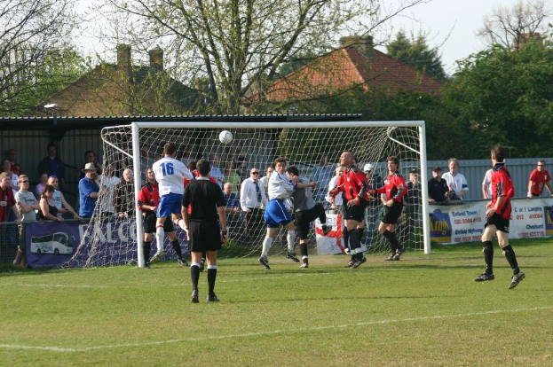 Spieler spielen Fußball auf einem Feld mit einem Tornetz, während Zuschauer dahinter stehen; Bäume und Häuser sind im Hintergrund sichtbar.