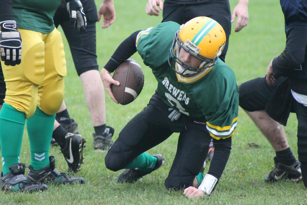 Ein Mann in einem grünen T-Shirt und einem gelben Helm hält einen American Football auf einem grasbewachsenen Feld, auf dem viele Menschen Fußball spielen.