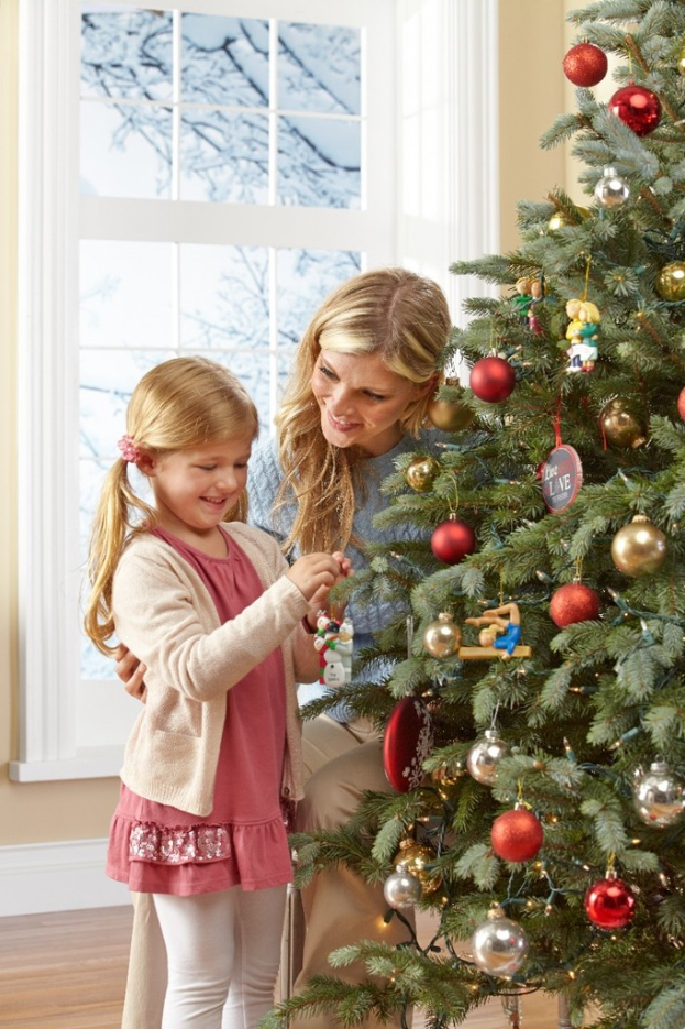 Ein geschmückter Weihnachtsbaum steht in einem Haus mit einer Frau und einem Kind im Vordergrund, einem Fenster und einer Wand im Hintergrund und Bäumen außerhalb des Fensters.