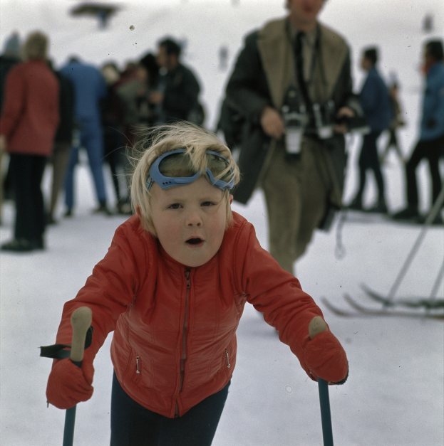 Ein junger Junge in roter Jacke und Brille fährt Ski auf einem schneebedeckten Hügel, hält Ski-Stöcke in den Händen, mit mehreren Menschen und einigen mit Ski-Brettern im Hintergrund.
