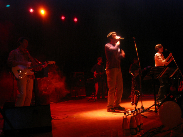 Zwei Personen singen in ein Mikrofon auf einer Bühne mit drei Männern, die Gitarren spielen und Wasserflaschen auf dem Podium.