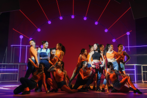 Eine Gruppe junger Tänzer in bunten Kostümen tanzt auf einer Bühne mit Geländern, beleuchtet von bunten Lichtern, vor einem jubelnden Publikum.