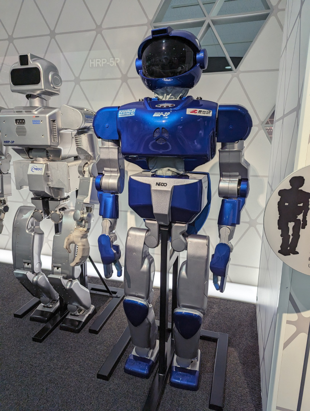 Zwei humanoide Roboter stehen nebeneinander in einem Raum mit einer Tafel auf der rechten Seite und einer Wand im Hintergrund, wahrscheinlich Teil einer Robotik-Ausstellung.