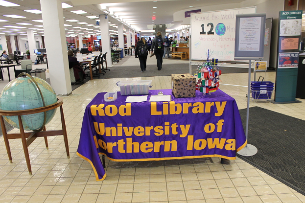 Eine Ausstellung mit einem Tisch, auf dem verschiedene Landesflaggen, eine Box, Papiere und ein Banner liegen, eine Kugel links daneben, ein Anschlagbrett mit einigen Leuten rechts daneben und mehreren Tischen und Stühlen links.