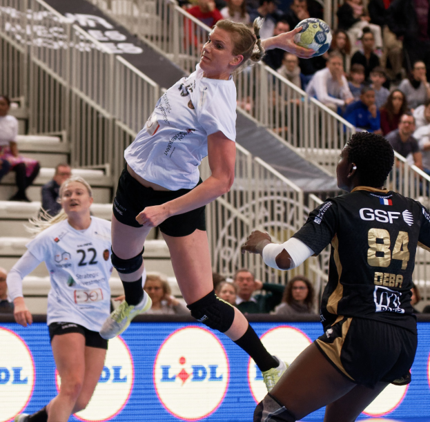 Eine Gruppe von Frauen, die Handball auf einem Platz spielen, mit einem Ball in der Luft, Zuschauern im Hintergrund und einer "Handball-Weltmeisterschaft 2019 - Tag 2"-Tafel.