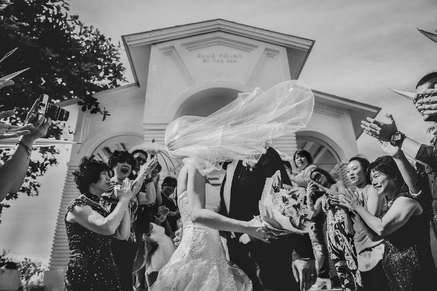 Ein Brautpaar steht in der Mitte des Bildes, mit einem Gebäude im Hintergrund, einer Person, die eine Kamera auf der linken Seite hält, einem Baum und einem Bukett, unter einem Himmel.