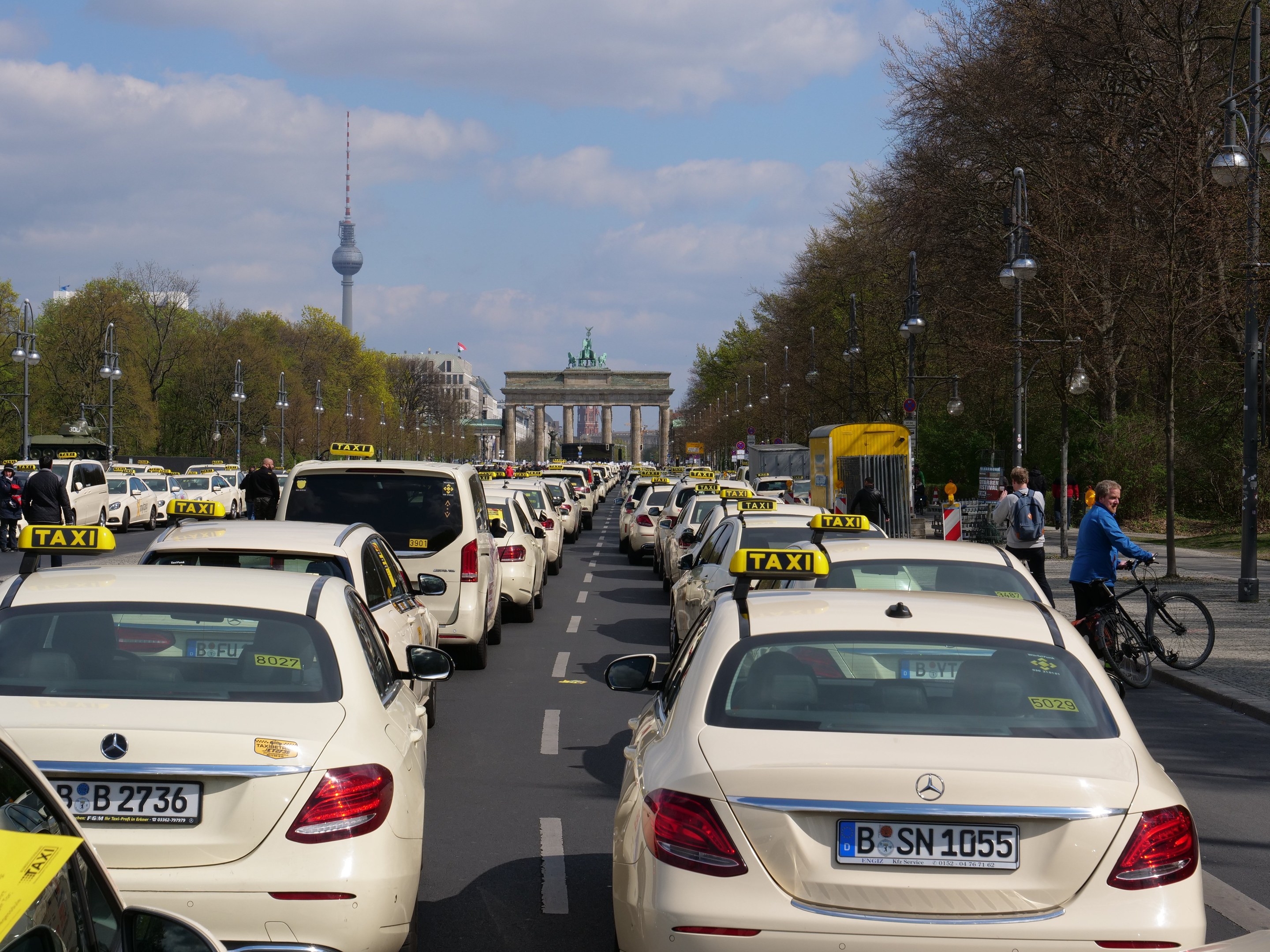 Eine lange Reihe von Taxis, die an einer belebten Straße in Berlin, Deutschland, geparkt sind, mit Fahrradfahrern und Fußgängern auf dem Gehweg, flankiert von Laternenpfählen und Bäumen, und Gebäuden, einem Bogen und einem Turm im Hintergrund unter einem bewölkten Himmel.