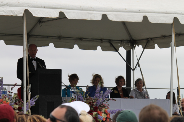 Eine Gruppe von Menschen an einem Tisch mit Blumen und Flaschen, eine Person in der N√§he eines Podiums mit einem Mikrofon auf einem St√§nder, unter einem Pavillon mit sichtbaren Pf√∂len.